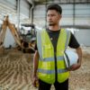 Portrait of a construction worker in safety gear holding a helmet inside an industrial setting.