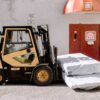 Forklift unloading wrapped goods outside an industrial building with a red door.